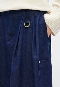 Donkerblauwe spijkerbroek met een wijde pijp, voorplooien, een gouden metalen ringdetail en een zijzak met een metalen knoopaccent.