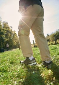Outdoor hiking shoes in black and olive green with a textured upper. Tan pants and white socks visible while walking on grassy path.