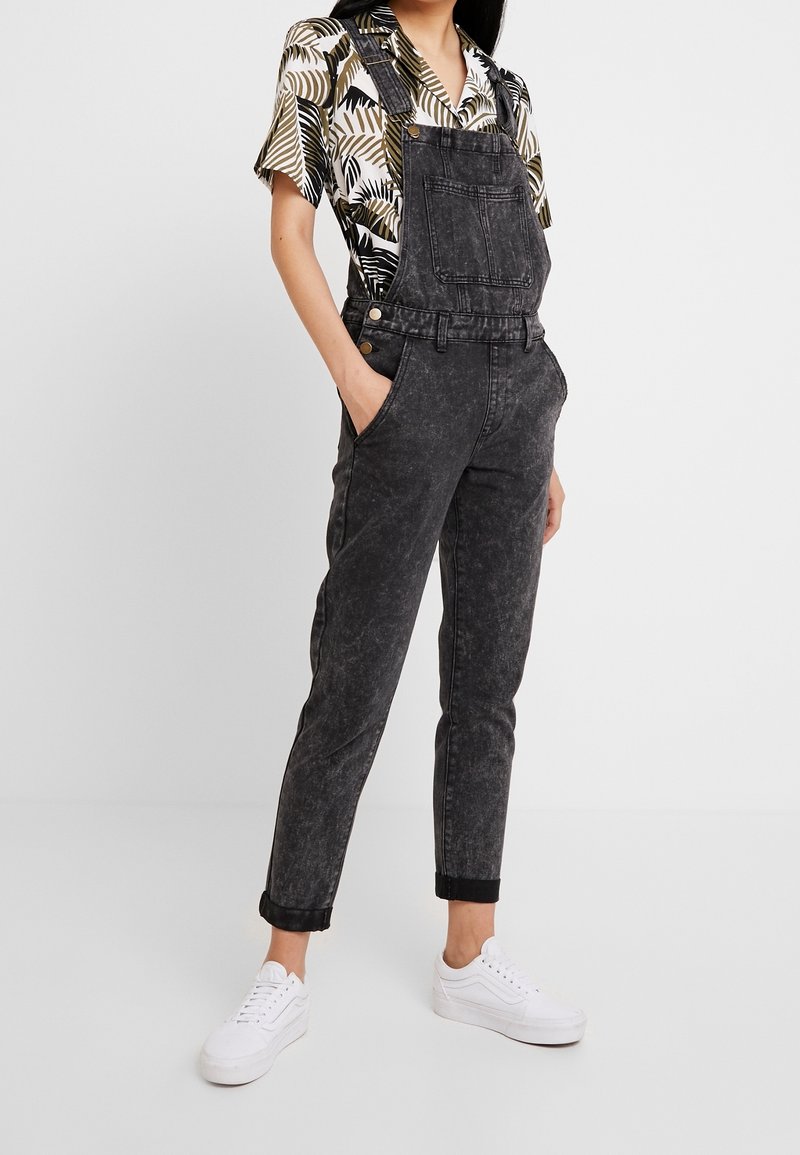 Salopette en denim noir avec une coupe ample, des bretelles ajustables et des poches avant. Portée avec une chemise à manches courtes à motifs et des baskets blanches.