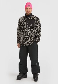 Veste en polaire avec un motif abstrait noir et blanc, poche avant, portée avec un pantalon noir et des bottes de ski noires. Bonnet en tricot rose sur la tête.