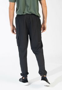 Zwarte joggerpants met een elastische tailleband en boorden, voorzien van een zijcargo zak en een zachte, lichte stof. Draagt met zwarte sneakers.