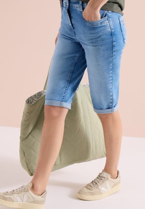 Personne portant un short en denim bleu clair retroussé, des baskets beiges, tenant un grand sac matelassé vert olive sur un fond uni.