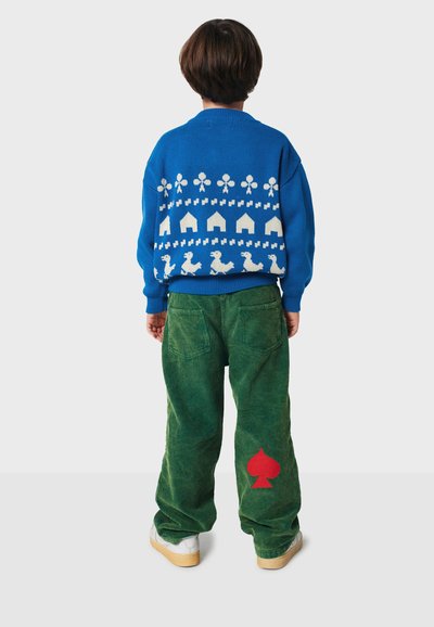 Pletený modrý sveter s bielymi vzormi, skombinovaný so zelenými kordovým nohavicami s červeným dizajnom srdca a bielymi teniskami.