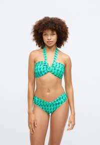 Zaļš bikini komplekts ar ģeometriskām zīmēm. Augšdaļai ir griezums ar pagrieztu dizainu un halteratām siksnām, savukārt apakšdaļa ir ar vidēju augstumu.