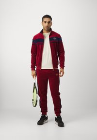 Sarkana samta treniņtērps ar tumši zilo svītru, ar rāvējslēdzēju aizdares jaku un atbilstošām biksēm. Iespējamās iezīmes ir logo un tenisa racket rokā. Melni sporta apavi.