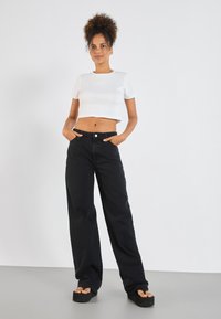 Fiatal nő göndör hajjal, fehér cropped toppan, fekete bő szárú nadrágban és fekete platform szandálban áll, kezét a zsebében tartva.