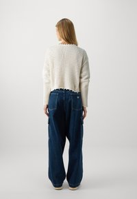 Maglione bianco lavorato a maglia con vestibilità ampia, caratterizzato da un motivo testurizzato e orlo sfilacciato; abbinato a jeans cargo blu scuro a gamba larga con tasche.