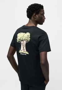 Μαύρο βαμβακερό T-shirt με ανοιχτοπράσινο καρτούν δέντρο στην πλάτη και κείμενο «ΦΙΛΟΙ ΤΗΣ ΦΥΣΗΣ» στο κάτω μέρος. Κοντά μανίκια, στρογγυλή λαιμόκοψη.