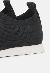 Svarte strikkede sneakers med teksturert overdel, elastiske mansjetter og hvit gummisåle. Har et sømløst design og ingen synlige snøring.