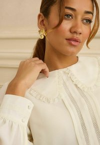 Világosbarna hajú nő, arany virág alakú fülbevalóval, fehér blúzzal, amelynek csipkés Peter Pan gallérja és díszes mandzsettái vannak.