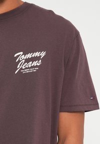 Lilla puuvillane T-särk ümmarguse kaelusega, millel on valge "Tommy Jeans" logo ja tekst, lühikesed varrukad ning väike logo lipp allääres.