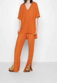 Henkilö, joka on pukeutunut löysään ribattu oranssiin V-päähineeseen ja siihen sopiviin leveälahkeisiin housuihin, joissa on edessä halkioita, yhdistettynä vaaleisiin läppäkenkiin, joissa on niittejä.