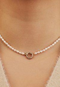 Collier ras du cou en perles de culture, composé de petites perles blanches polies et d'un fermoir circulaire doré central avec des accents en boucle.