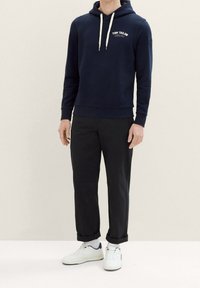 Marinblå hoodie med dragsnöre och logotyp på bröstet, matchad med svarta byxor och vita sneakers. Mjuk tyg och avslappnad passform.