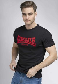 Fekete, rövid ujjú póló piros "LONSDALE" logóval és "LONDON" felirattal. Az anyag puhanak tűnik, és a szabása testhez simuló.