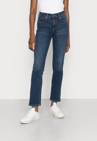 Vaqueros de denim azul con corte ajustado, cintura media, diseño de cinco bolsillos y ligera elasticidad. Combinados con zapatillas blancas, sobre un fondo neutro.