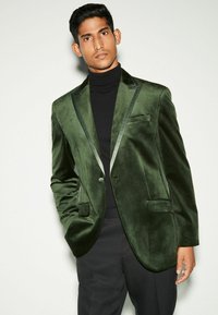 Samtgrünes Blazer mit Satinbesatz, ein Knopfverschluss und Seitentaschen, über einem schwarzen Rollkragenpullover getragen, vor einem neutralen Hintergrund präsentiert.