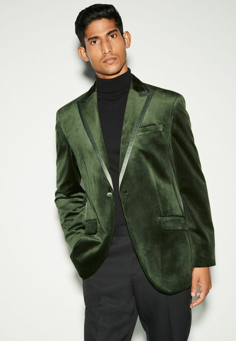 Samtgrünes Blazer mit Satinbesatz, ein Knopfverschluss und Seitentaschen, über einem schwarzen Rollkragenpullover getragen, vor einem neutralen Hintergrund präsentiert.