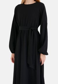 Musta mekko pitkillä puff-hihnoilla, solmittu vyötärö ja rento leikkaus. Valmistettu kevyestä kankaasta, jossa on hienovarainen rakenne ja pyöreä kaula-aukko.