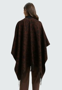 Frau mit langem braunem Haar, die einen dunkelbraunen Poncho mit Tierdruck und Fransen am Saum trägt, steht mit dem Rücken zum Betrachter vor einem schlichten Hintergrund.