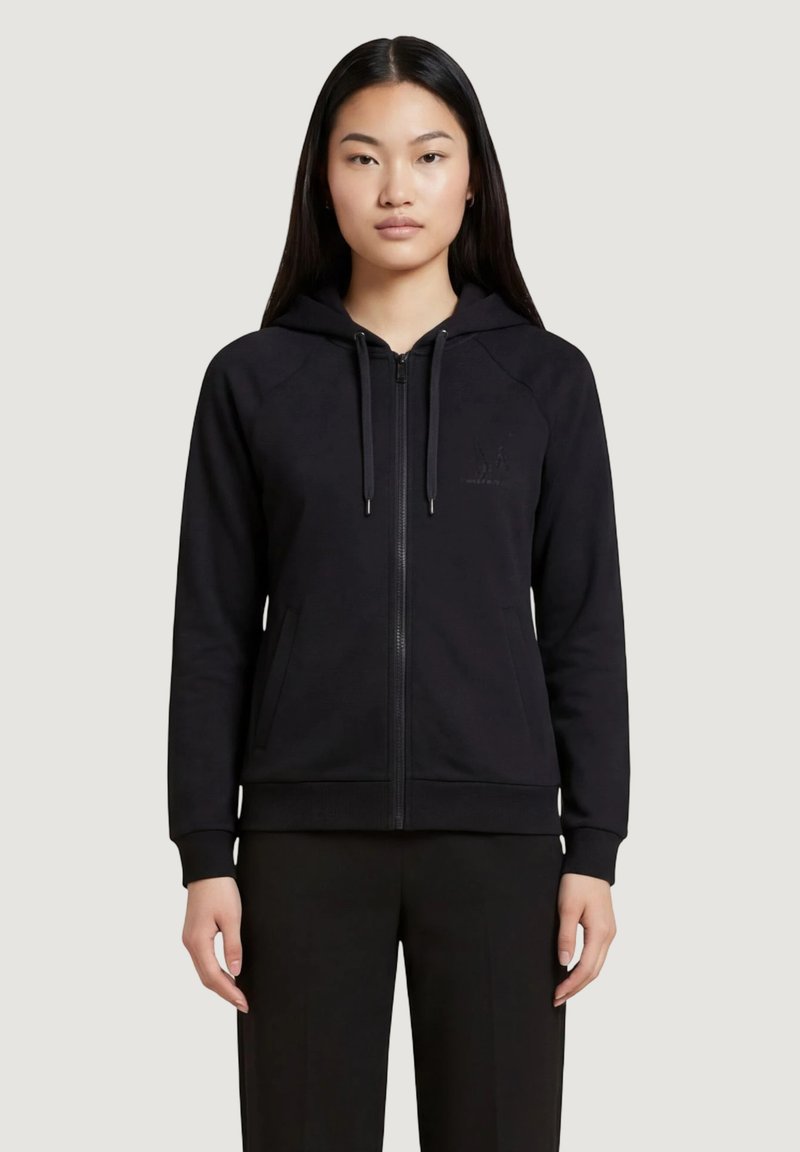 Femme aux longs cheveux noirs portant un sweat à capuche noir zippé et un pantalon noir, debout devant un fond clair uni.