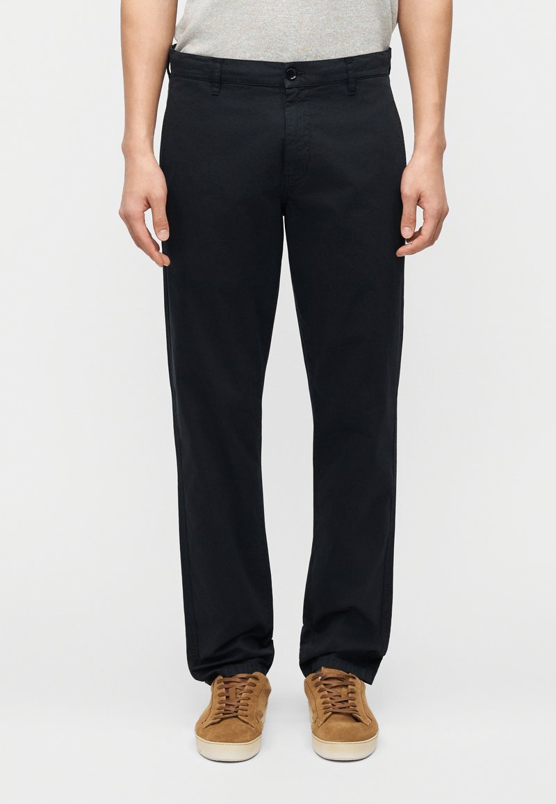 Pantalon noir à jambes droites en coton avec une texture lisse, doté d'une fermeture avant à boutons et de passants pour ceinture. Porté avec des baskets marron.