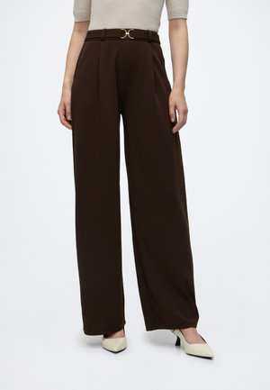 Woman wearing high-waist dark brown wide-leg trousers with belt and cream pointed-toe heels against a plain background.