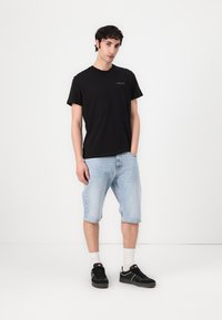 Schwarzes T-Shirt mit kurzen Ärmeln und aufgedrucktem Logo. Hellblaue Jeansshorts, bis zur Mitte des Oberschenkels. Schwarze Sneakers mit weißen Sohlen und knöchellangen weißen Socken.