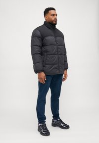 Sort pufferjakke med høj krave, quiltet design og elastiske manchetter, parret med mørkeblå jeans og sorte sneakers.