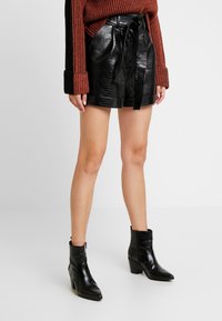 Černé šortky z umělé kůže s krokodýlí texturou, s vázáním v pase, spárované s černými kotníkovými botami a svetr s pleteninou v cihlové barvě.