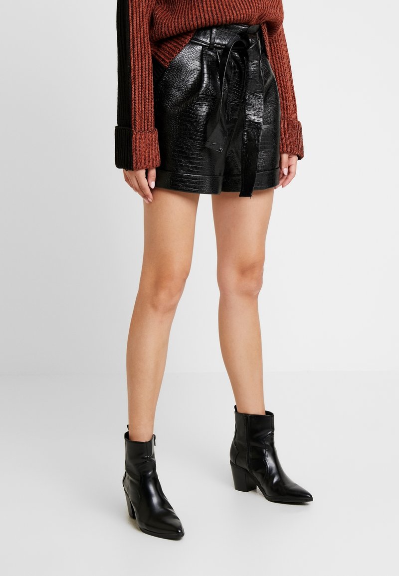 Černé šortky z umělé kůže s krokodýlí texturou, s vázáním v pase, spárované s černými kotníkovými botami a svetr s pleteninou v cihlové barvě.