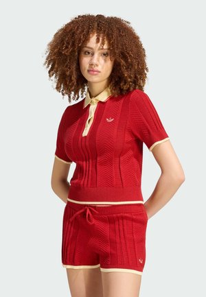Femme aux cheveux bouclés portant un haut en maille rouge à manches courtes avec col beige et un short en maille rouge assorti avec bordure beige, debout les mains dans le dos.