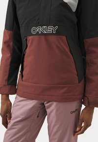 Veste isolante noire et maroon mettant en avant un logo Oakley, avec des poches zippées et un tissu lisse et durable. Associée à un pantalon rose.