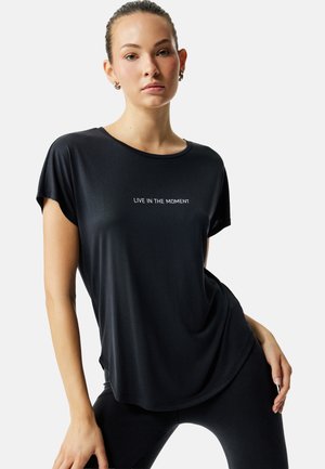 Ung kvinde med håret bundet tilbage iført en sort skjorte med teksten "LEV I ØJEBLIKKET" i hvid, poserende med hånden på hoften mod en hvid baggrund.
