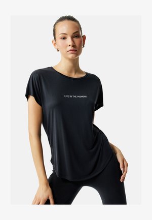 Ung kvinde med håret bundet tilbage iført en sort skjorte med teksten "LEV I ØJEBLIKKET" i hvid, poserende med hånden på hoften mod en hvid baggrund.