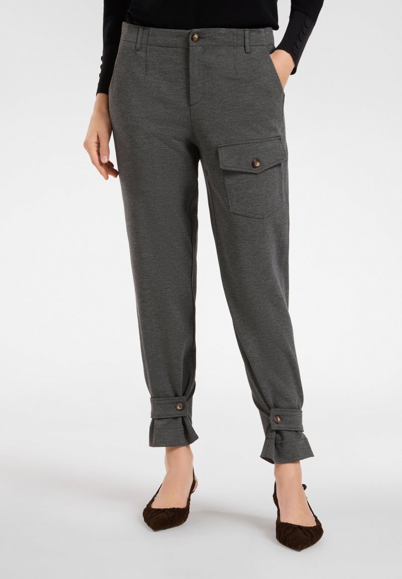 Kobieta ubraną w ciemnoszare zwężane spodnie z mankietami zapinanymi na guziki oraz boczną kieszenią z klapą, w zestawieniu z ciemnobrązowymi balerinami z ostrym noskiem.