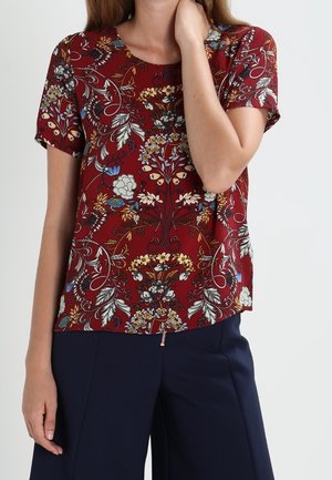 Vrouw die een kortemouwen bloes draagt in bordeauxrood met bloemmotief en marineblauwe wijde broek, poseert met één hand bij haar hals.