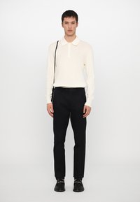 Camisa pólo de manga longa de cor creme com colarinho, combinada com calças pretas e sapatos pretos. Design minimalista, textura suave.