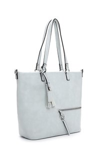 Sac fourre-tout gris clair avec double bandoulière, poche zippée à l'avant, et un pompon décoratif suspendu près du haut.