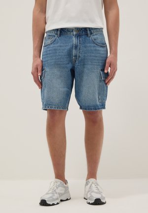 Uomo che indossa pantaloncini cargo in denim blu di media lunghezza con tasche a patta, sneakers bianche e una T-shirt bianca, in piedi su uno sfondo chiaro.