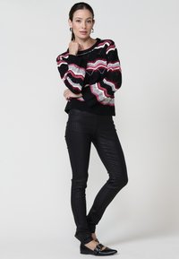 Pull noir avec des motifs en zigzag roses, blancs et rouges ; associé à un pantalon à carreaux noir près du corps et des chaussures plates noires.