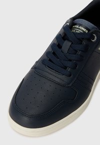 Nærbilde av en mørkeblå sneaker med teksturert lær, med rund tå, snøring og perforerte detaljer nær fronten.