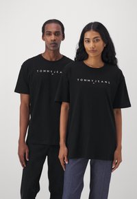 T-shirt in cotone nero con maniche corte, con il logo "TOMMY JEANS" in bianco e rosso sul petto. I modelli indossano jeans e pantaloni neri.