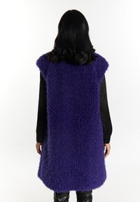 Violetti pörröinen liivi, jossa on hihaton muotoilu, pyöreä kaulus ja teksturoitu, pehmeä kangas. Päällä musta paita.