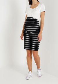 Graviditetsklänning med vit överdel och svartvit randig nederdel. Har en figurnära design och korta ärmar. Matchas med vita sneakers.