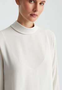 Witte blouse met een hoge, ronde hals en lange mouwen. Glad, lichtgewicht materiaal met een subtiele glans. V-vormige halslijn detail.