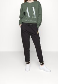 Zelená mikina s bílým logem "AX", kombinovaná s černými tepláky s páskem na stažení a zlatými detaily, nošena s bílými teniskami.