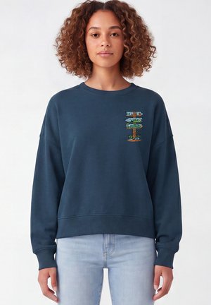 Vrouw met krullend haar draagt een donkerblauwe sweatshirt met een kleurrijk cartoonwegwijzerontwerp nabij de linker schouder en lichtblauwe spijkerbroek.