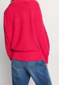 Pull tricoté texturé rose vif porté avec un jean en denim bleu, vu de dos, mettant en valeur la coupe du pull et le détail du tissu.