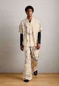 Beige, map-patterned short-sleeve shirt and loose pants set. Textured fabric with black accents. Model wears black long-sleeve top underneath.